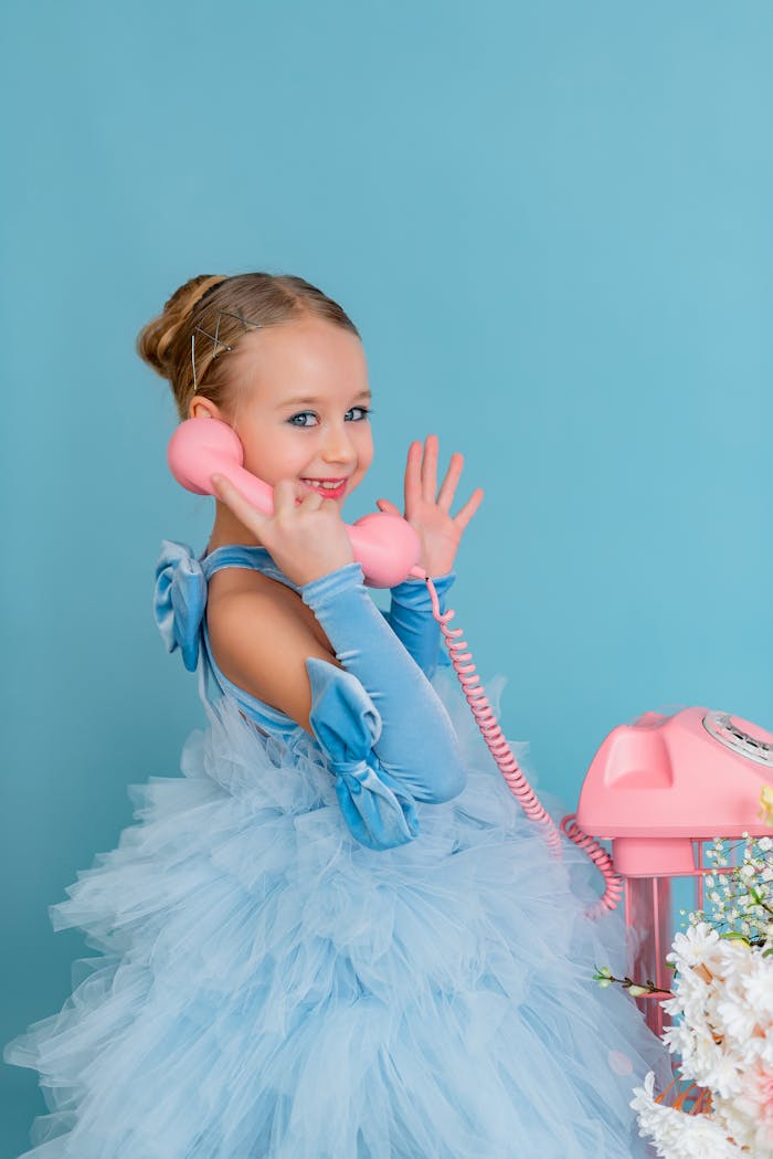 Adorable girl in blue gown holding a pink phone, smiling and waving in a studio setting.